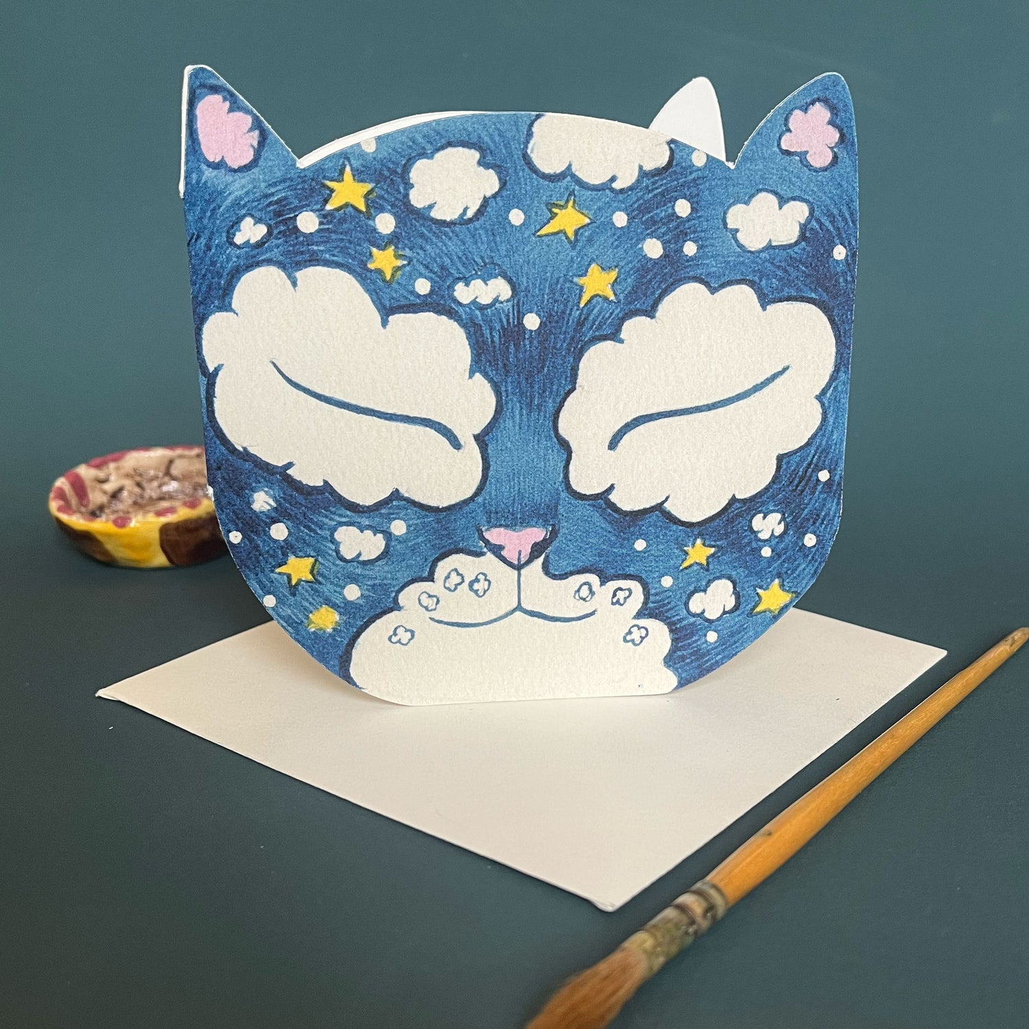 A die-cut card with a watercolor painting of a cat's face, featuring stars and a cloudy background, displayed on a table with artistic tools.