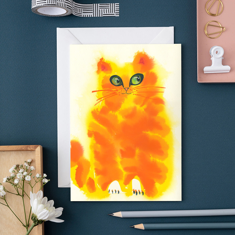 Greeting card with a colorful cat illustration on a blue surface with stationery items.
